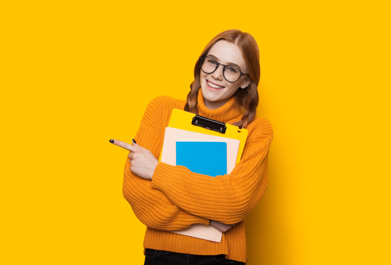 Monochrome portrait of a caucasian student with freckles and red hair posing with some books and eyeglasses on a yellow wall with free space