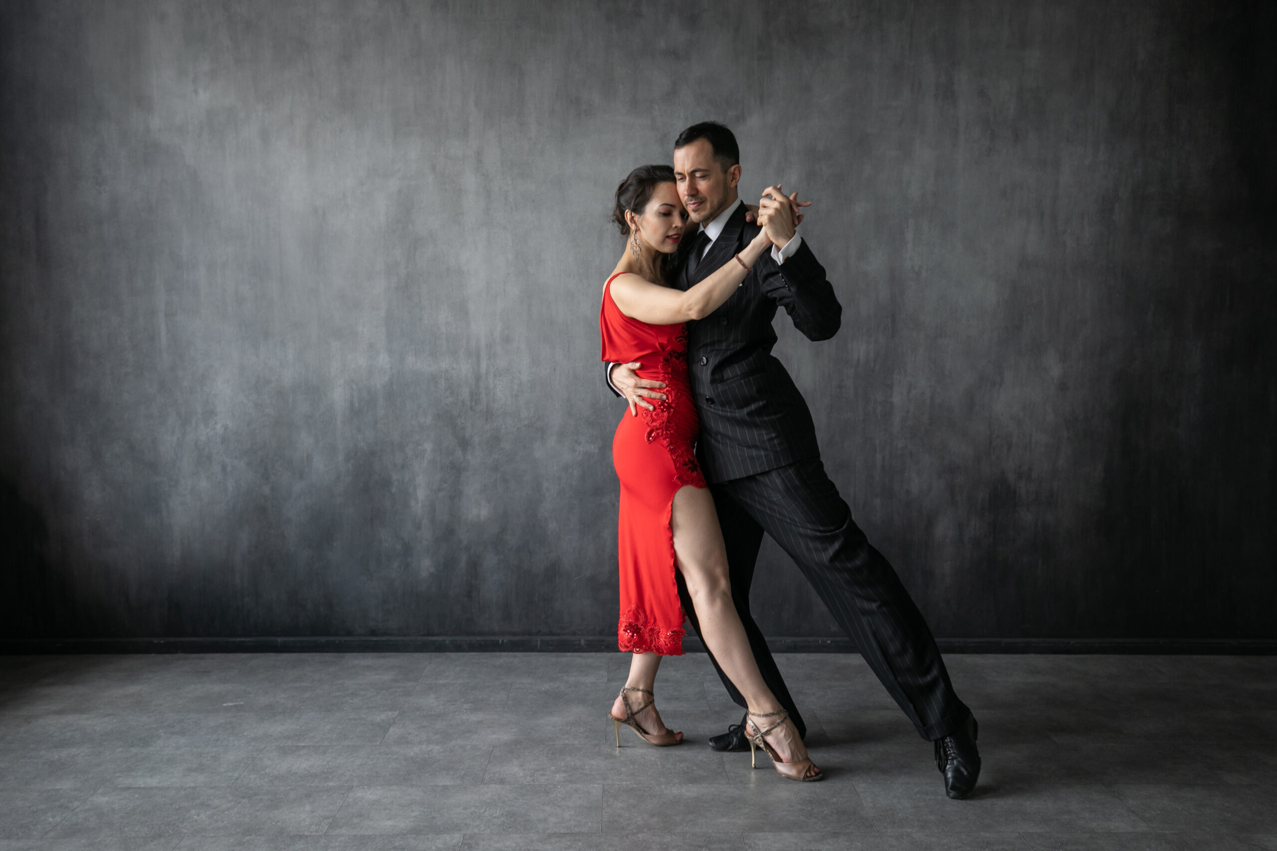 Couple of professional dancers in elegant suit and red dress in a tango dancing movement on dark background. Handsome man and woman dance cheek to cheek.
