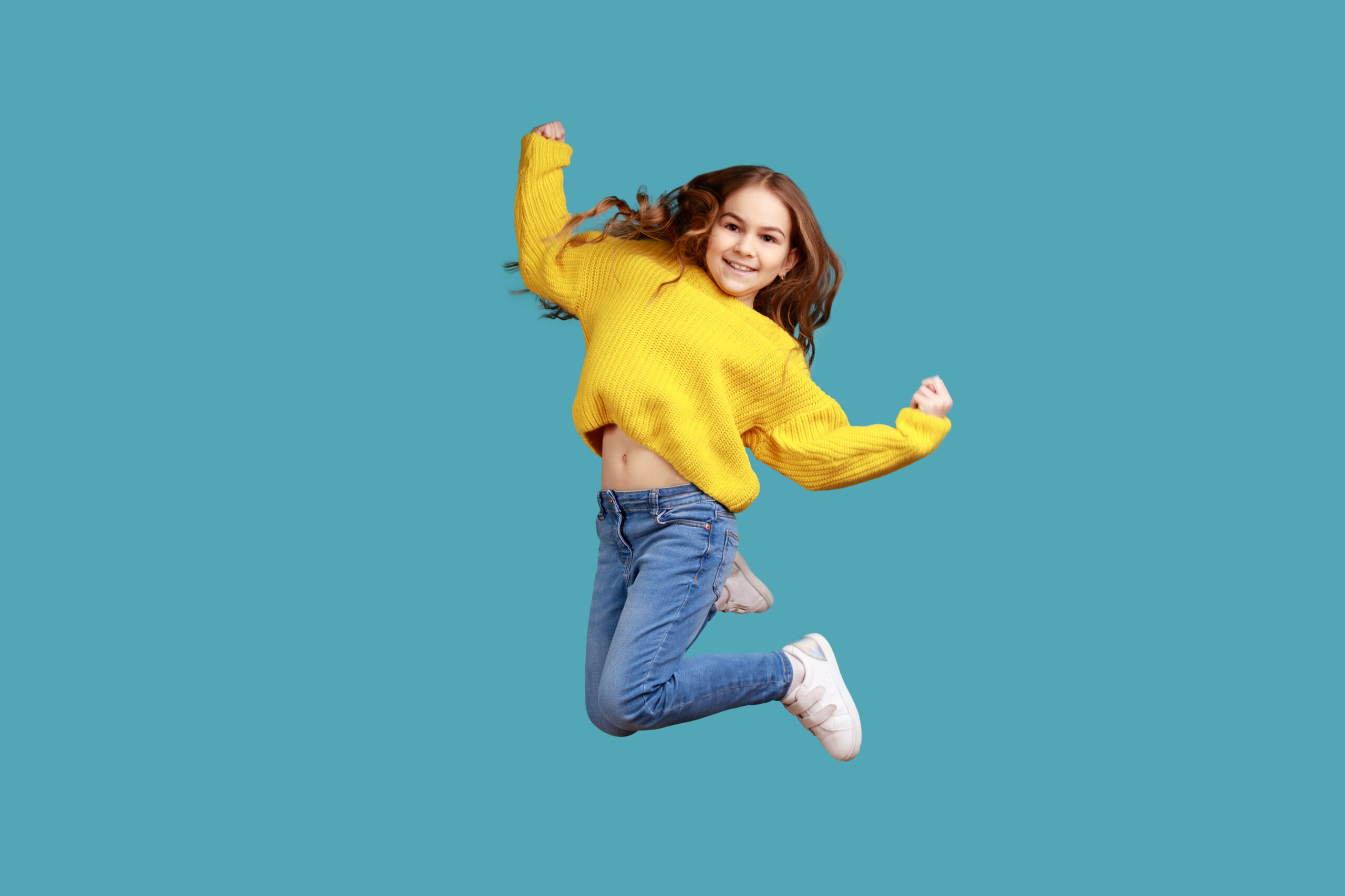 Full length portrait of little girl jumping up high, expressing happiness, celebrating success, wearing yellow casual style sweater. Indoor studio shot isolated on blue background.